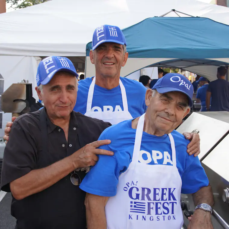 Three Greek Fest volunteers in OPA! caps and aprons sharing a laugh by the grill